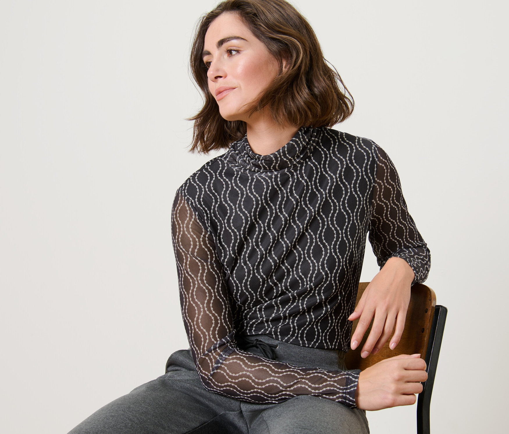 Egy nő ül egy széken, és mintás, garbónyakú mesh felsőt és szürke melegítőnadrágot visel.