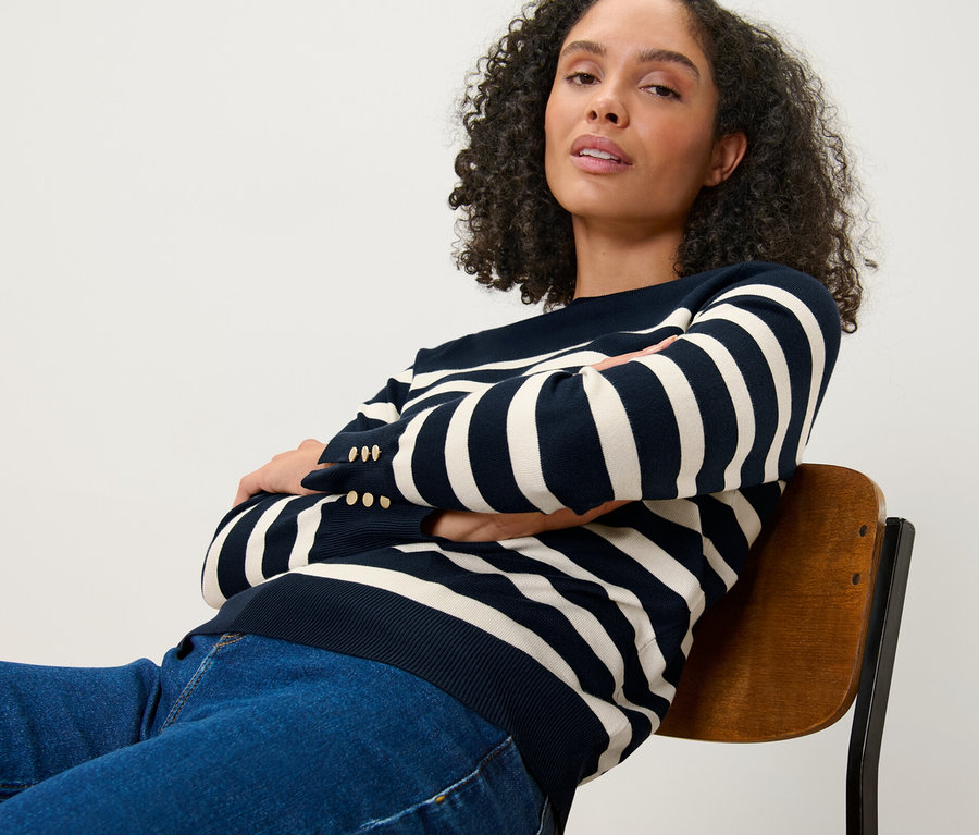 Nő ül széken keresztbe tett karokkal, sötétkék csíkos, finomkötésű pulóvert visel.