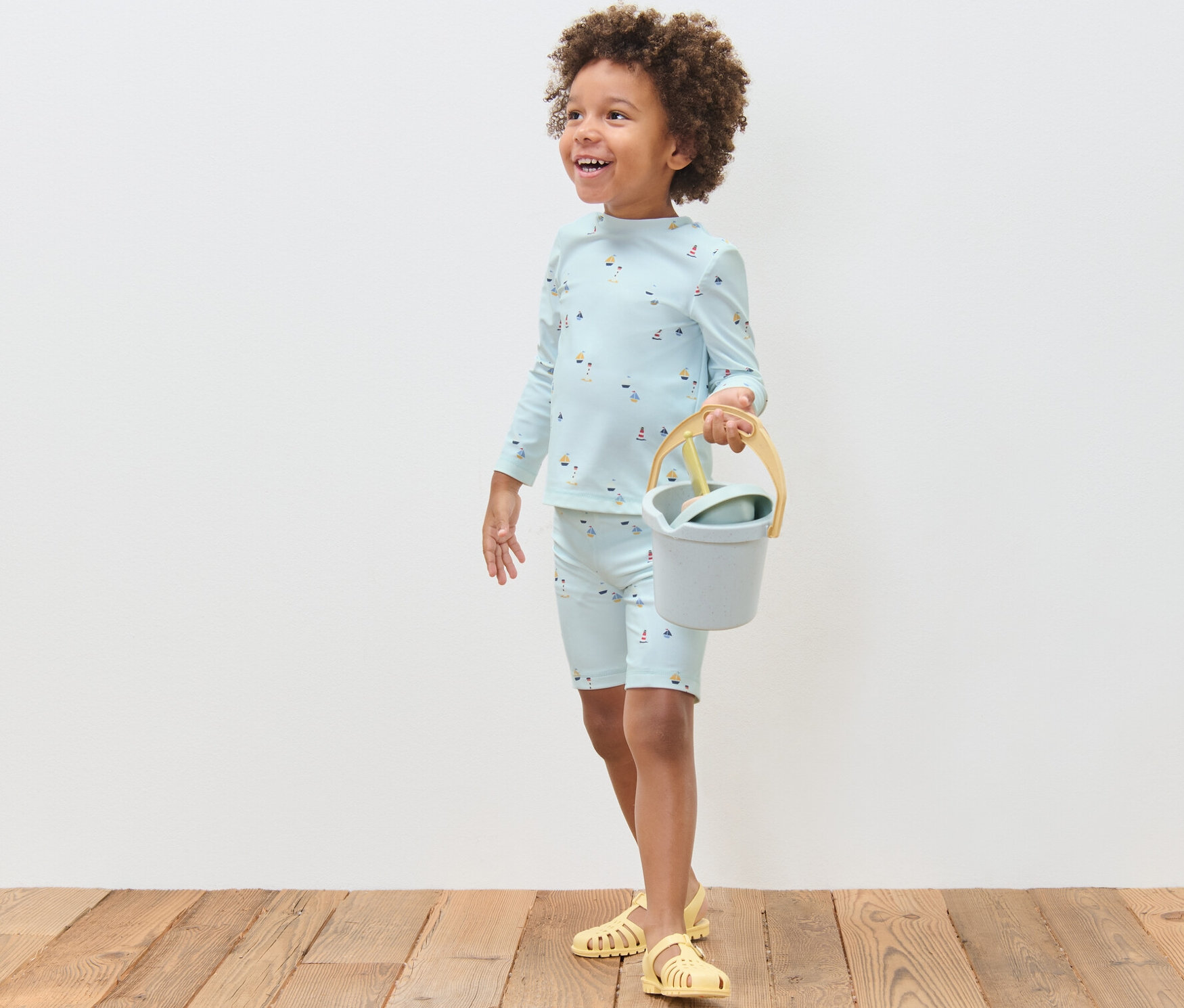 Fiúgyermek hajós mintás, UV-szűrős együttesben vödröt és lapátot tart.