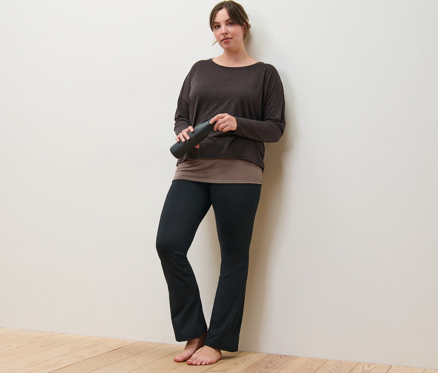 Egy nő a falnak dőlve tart egy barna 2 az 1-ben sportfelsőt és egy alakformáló sportnadrágot.