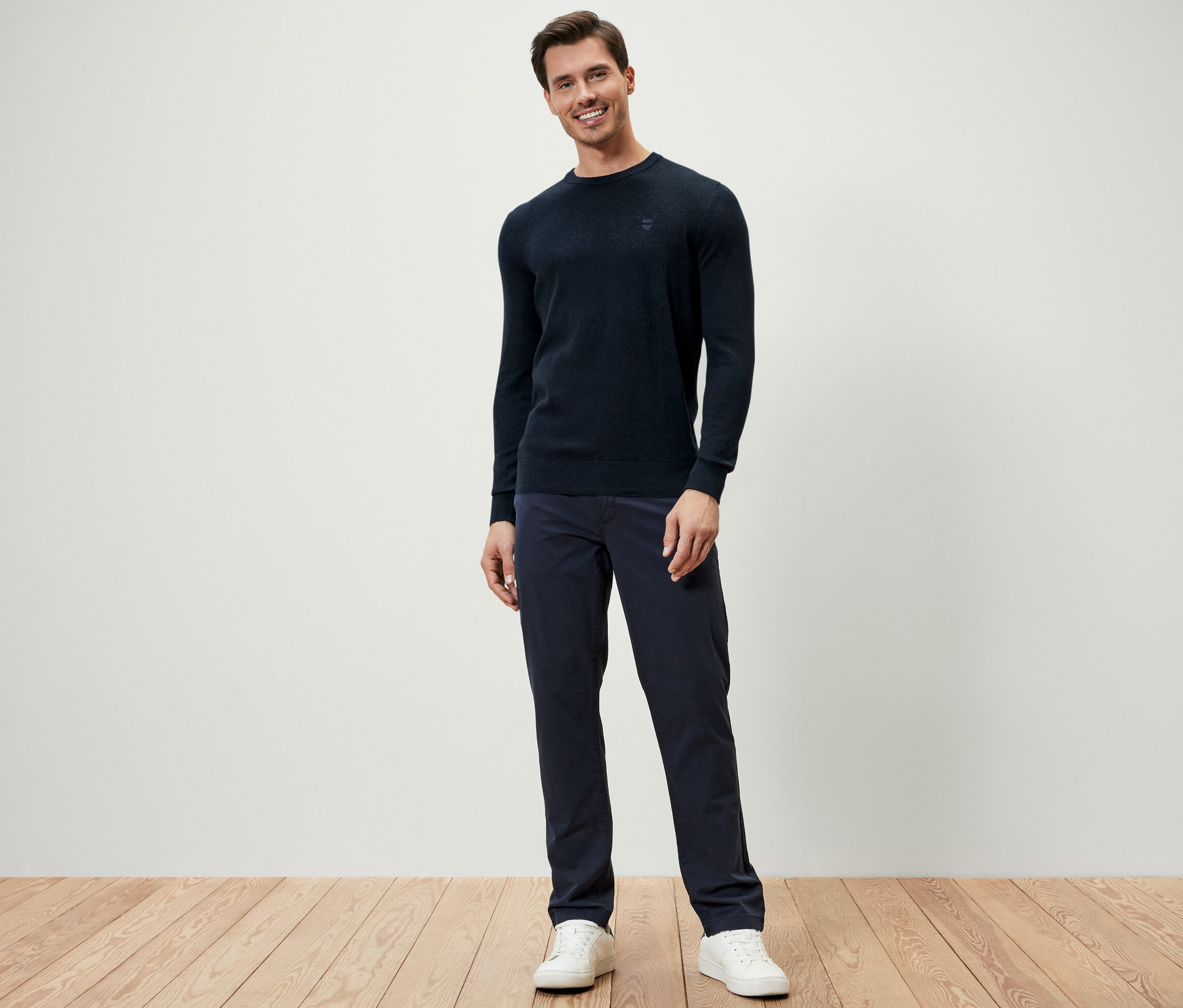 Egy férfi áll, sötétkék kötött pulóvert, sötétkék nadrágot és fehér tornacipőt visel.