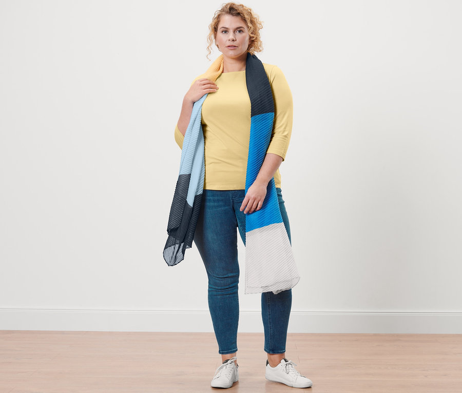 Nő pózol sárga, 3/4-es ujjú trikóban, farmer hatású treggingsben és kreppelt szövetsállal.