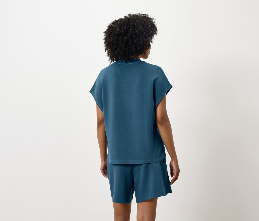 Egy nő áll háttal, kékeszöld felsőt és rövidnadrágot visel.