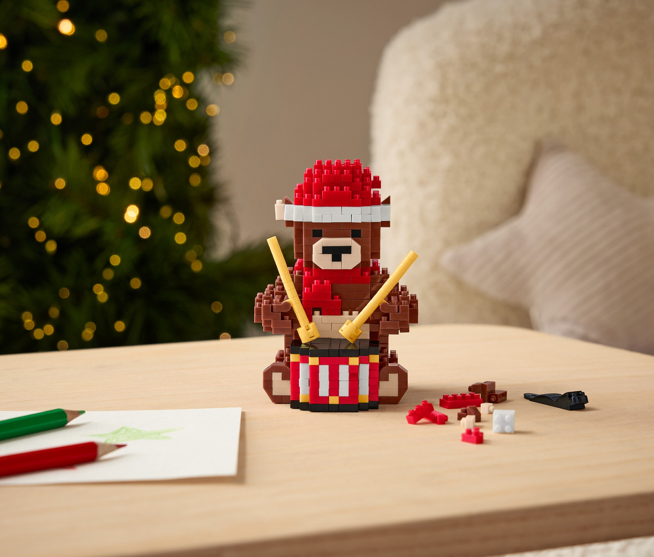 Egy kockákból álló medve piros sapkában és sálban dobol a pálcikákkal az asztalon.