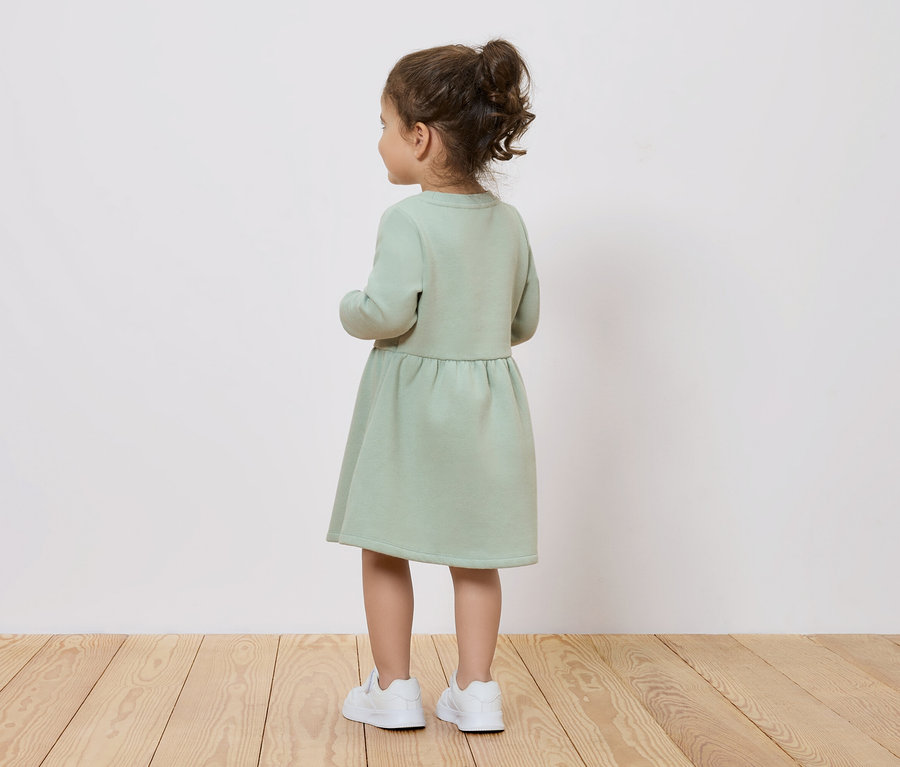 Kislány zöld ruhában és fehér cipőben, hátulról.