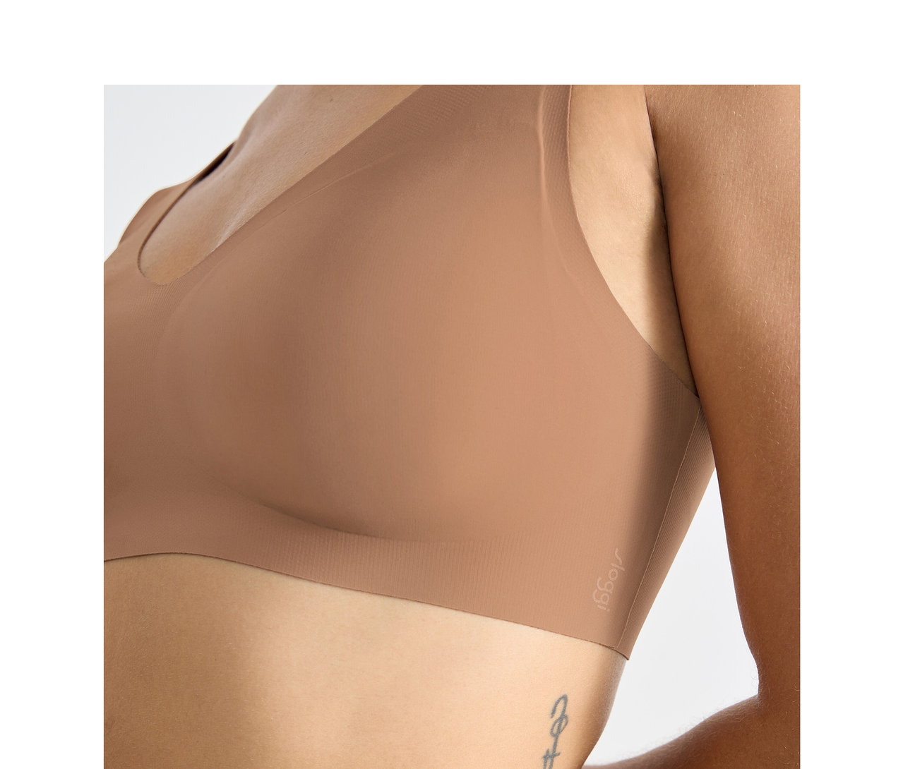 Egy személy közeli felvétele, aki sloggi ZERO Feel 2.0 Bralette melltartót visel nosztalgia barna színben, oldalán tetoválással.