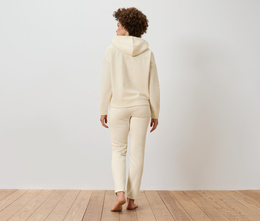 Egy göndör hajú nő háttal áll, bézs színű kapucnis pulóvert és jogging nadrágot visel a fapadlón.