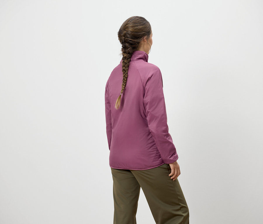 Egy copfos nő áll háttal, rózsaszín kabátot és zöld nadrágot visel.