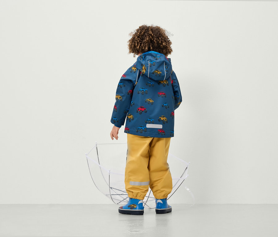 Egy fiú háttal a kamerának autó mintás kabátot és sárga nadrágot visel egy átlátszó esernyő mellett.