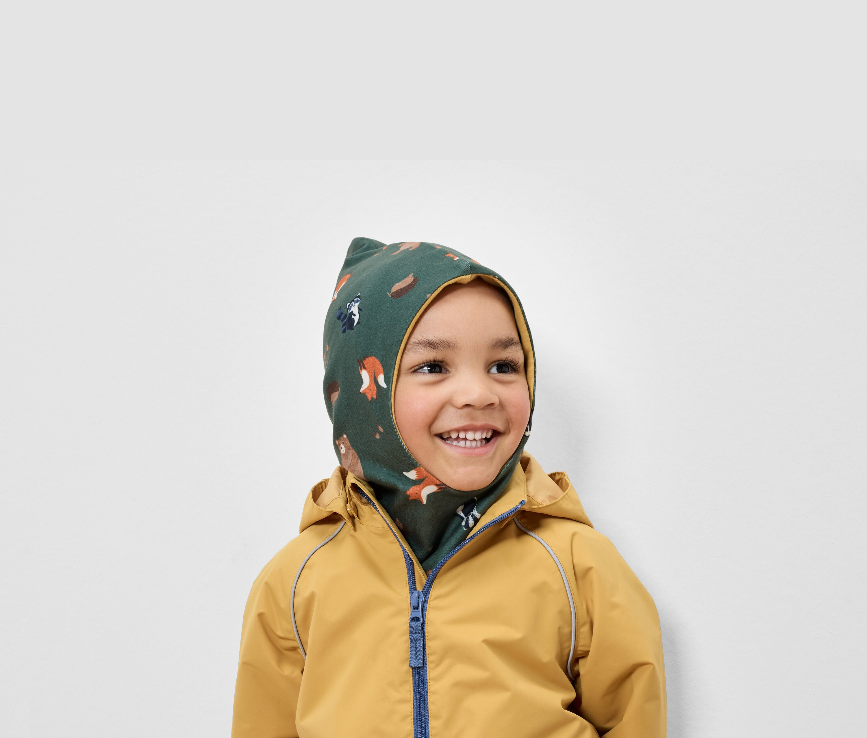 Mosolygó fiú zöld, állatmintás símaszkot és sárga kabátot visel.