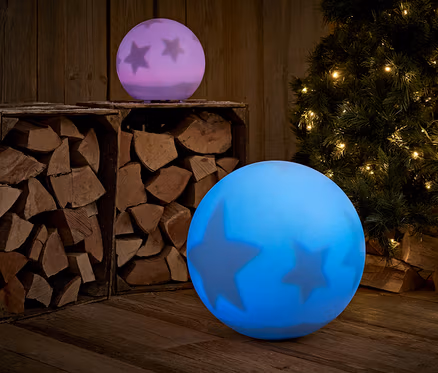 Két csillagokkal díszített világító gömb áll egy fapadlón, a háttérben fa és egy karácsonyfa.