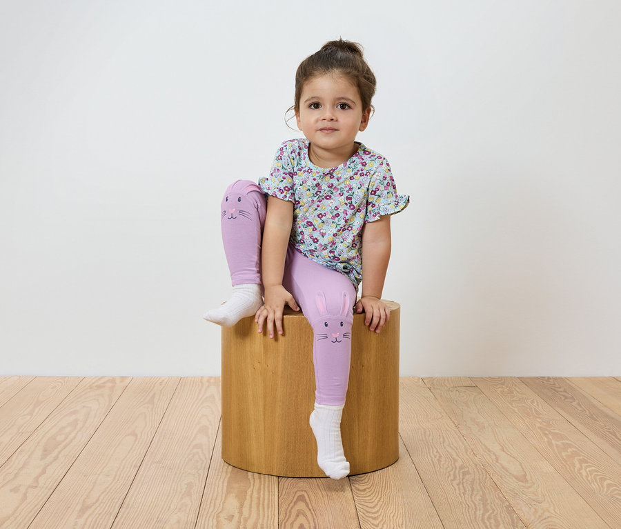 Egy kislány ül egy fa zsámolyon. Virágos pólót és lila, macskamintás leggingset visel.