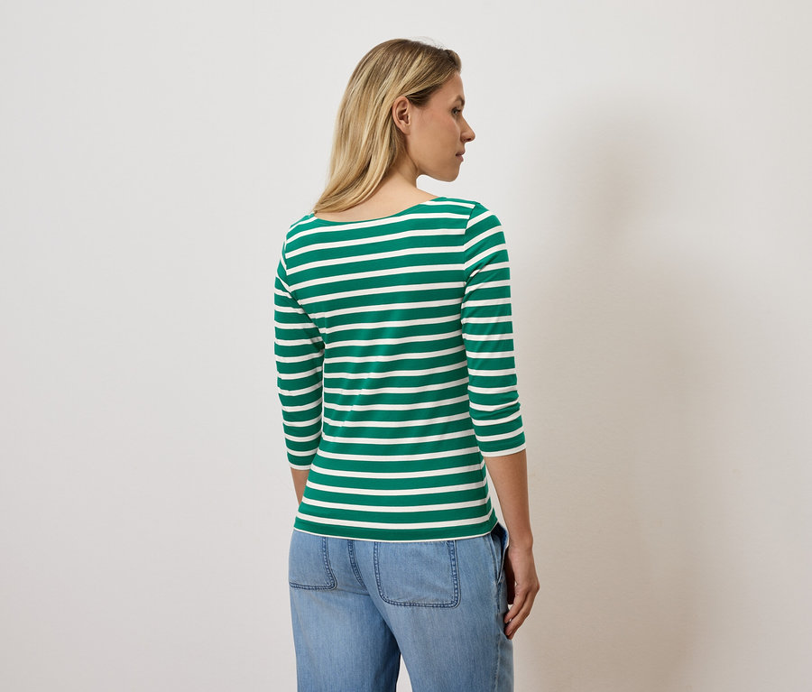 Egy szőke hajú nő háttal áll a kamerának, zöld-fehér csíkos, háromnegyedes ujjú pólót és kék farmert visel.