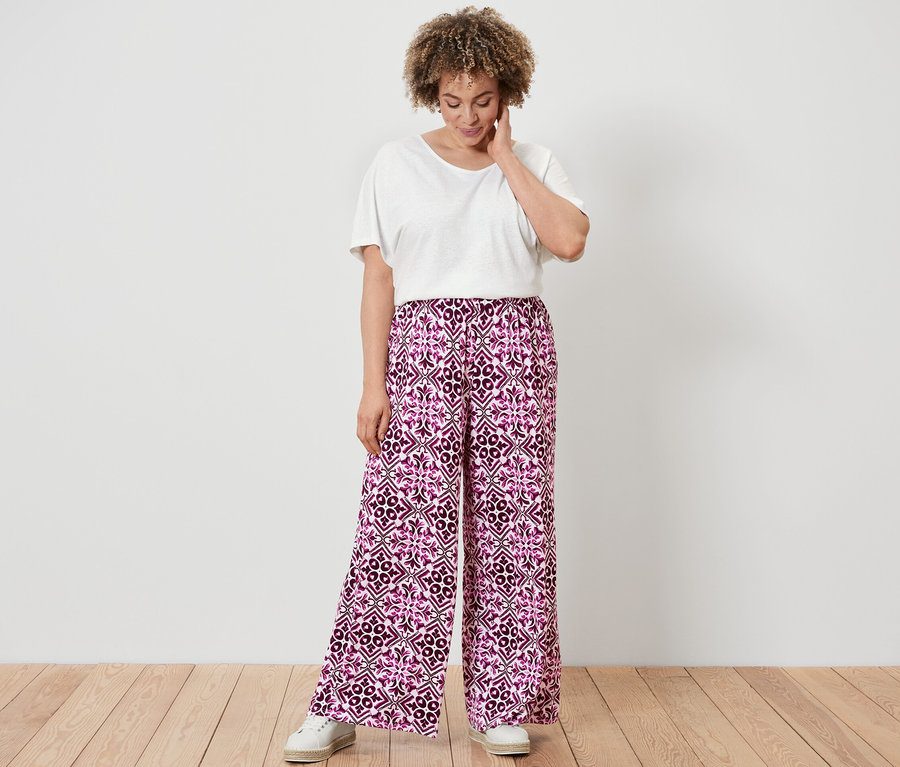 Egy nő fehér len-keverék pólóban és fukszia mintás, női bőszárú nadrágban áll egy fapadlón.
