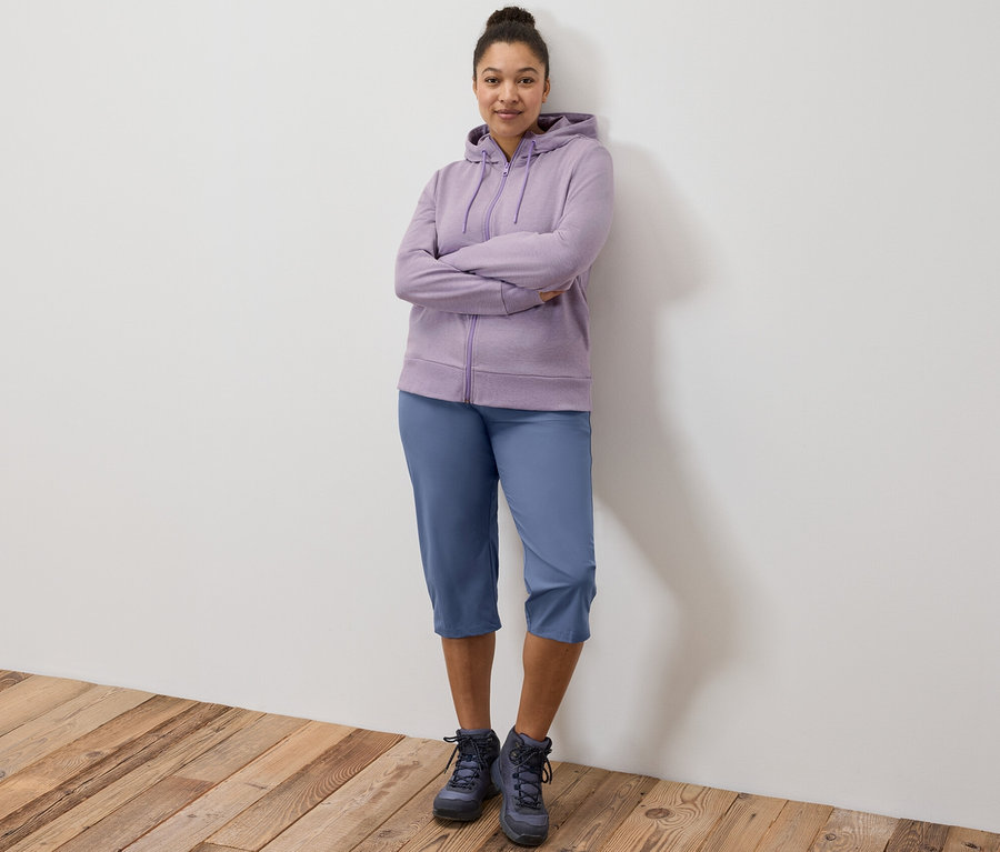 Egy nő áll keresztbe tett karokkal, lila szabadidőfelsőt és funkcionális térdnadrágot visel.