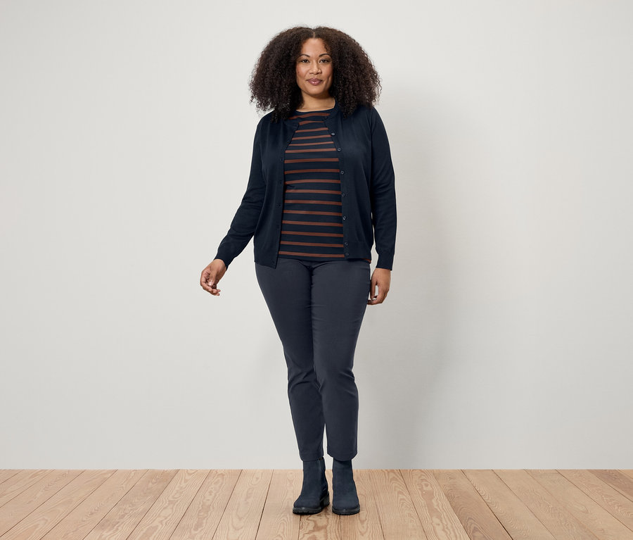 Sötét, göndör hajú nő pózol fapadlón, sötétkék kardigánt, csíkos pólót, sötétkék nadrágot és csizmát visel.