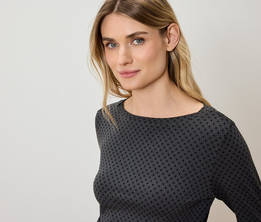 Portré egy szőke hajú, kék szemű nőről, aki egy szürke-fekete, geometrikus mintás felsőt visel.