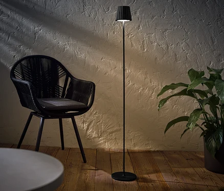 Egy fekete LED-es akkumulátoros kültéri állólámpa egy fekete fonott szék és egy cserepes növény mellett áll.