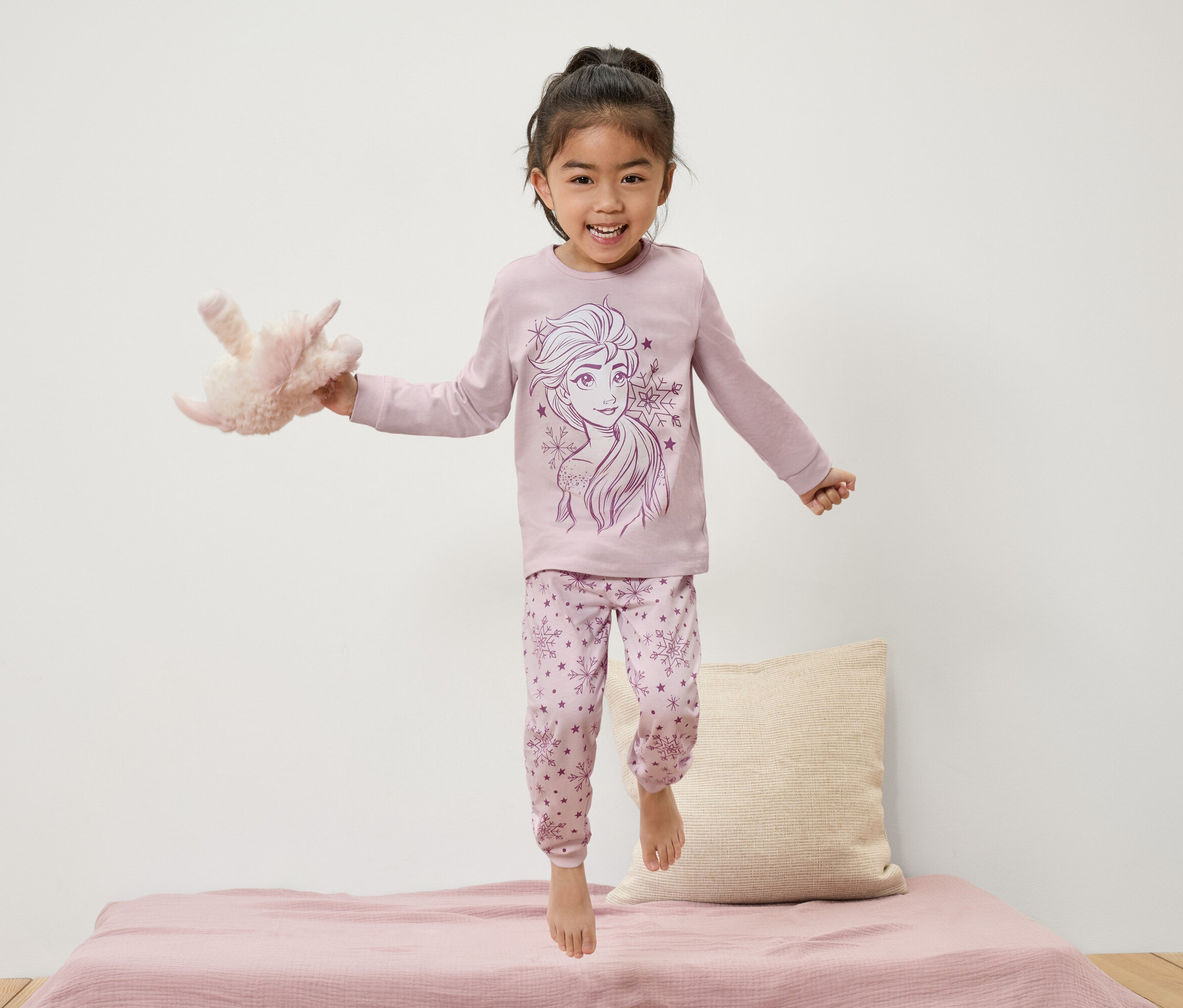 Elsa mintás pizsamában ugráló kislány az ágyon, plüssállatot tartva.
