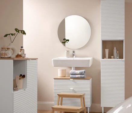 A fürdőszobában található egy Badhochschrank »SONARI« mit Türen, egy Waschbeckenunterschrank »SONARI« mit Schubladen és egy Badkommode »SONARI« mit Türen. Egy kb. 60 x 100 cm-es memóriahabos fürdőszobaszőnyeg található egy tömörfa Tritthocker aus Massivholz előtt. A mosdószekrényen két világoskék prémium törölköző található.