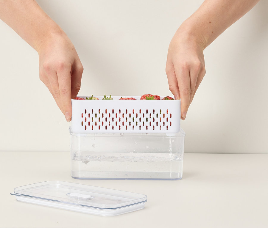 Kezek helyeznek egy fehér epret tartalmazó tartályt egy vízzel teli átlátszó tartályra.