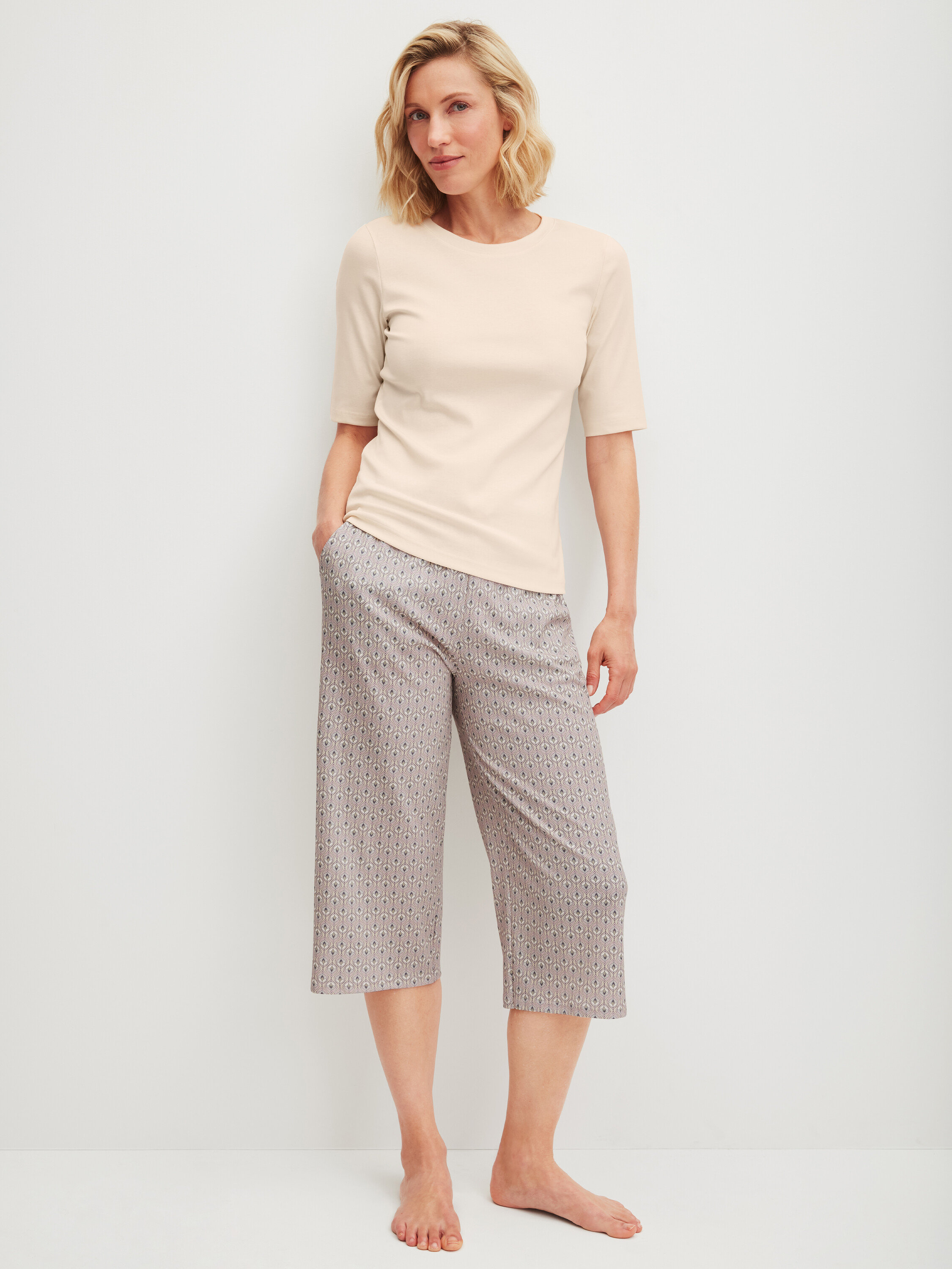 Egy nő áll mezítláb, világos pólót és mintás háromnegyedes nadrágot visel.