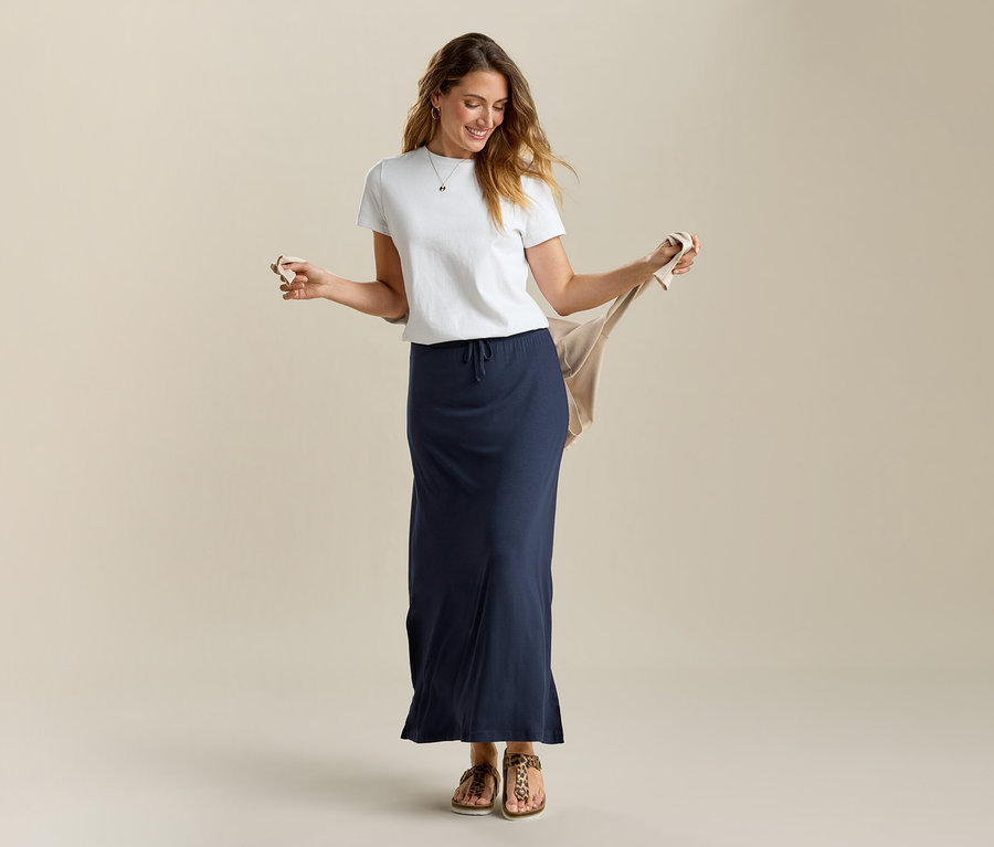 Nő bézs kardigánt tart, fehér pólót és sötétkék hosszú jersey szoknyát visel.