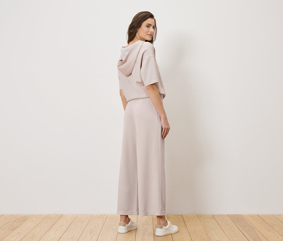 Egy nő világos rózsaszín nadrágkosztümben és fehér tornacipőben áll, fejét a válla fölé fordítva.