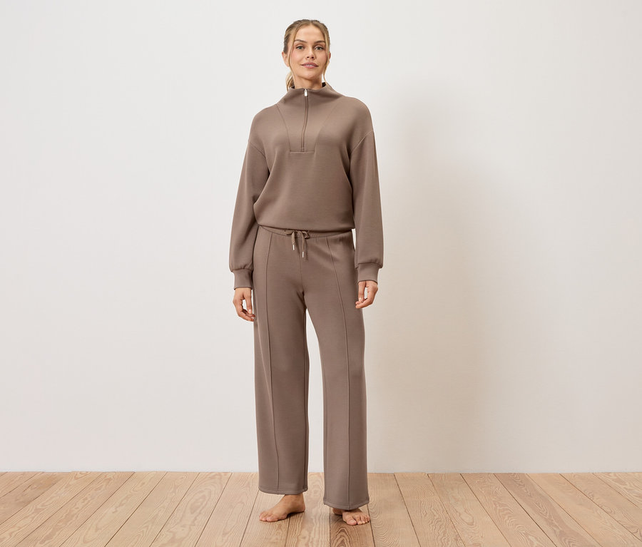 Nő áll mezítláb a fapadlón, barna tréningruhát visel.