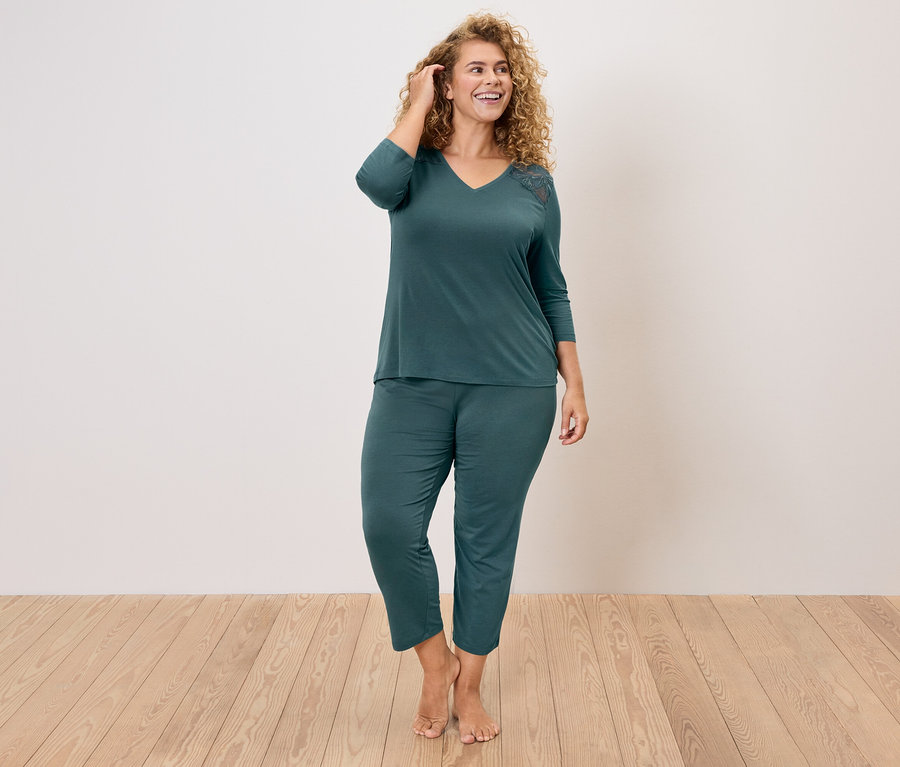 Göndör hajú nő, aki egy sötétzöld pizsama szettet visel, amely egy V-nyakú felsőből és egy csipke applikációból áll a vállán, valamint egy nadrágból.