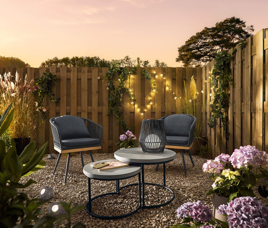 Két, textil keresztszőttel ellátott forgó kerti szék áll 2 db policement asztallapos kerti asztal mellett. Az egyiken egy kis, textilszalagos napelemes viharlámpa áll. Prémium napelemes LED-es kövek találhatók a kavicsban.