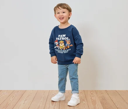Egy mosolygó fiú kék Paw Patrol pulóvert, farmert és fehér tornacipőt visel.