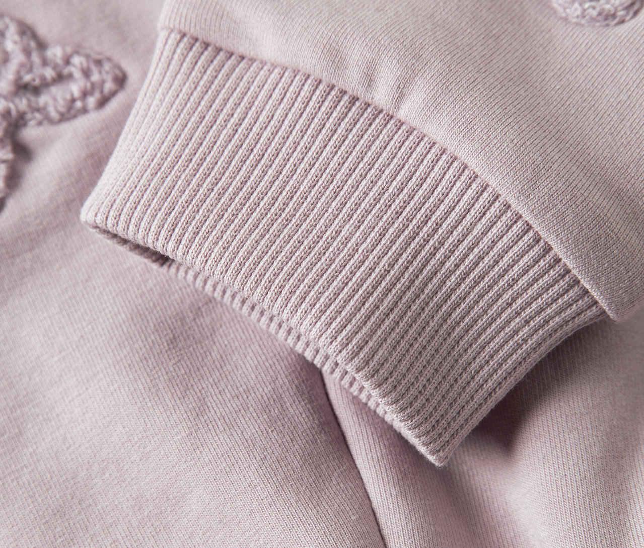 Egy lila színű pulóver ujjának közeli képe, bordázott szegéllyel és virágos applikációval.