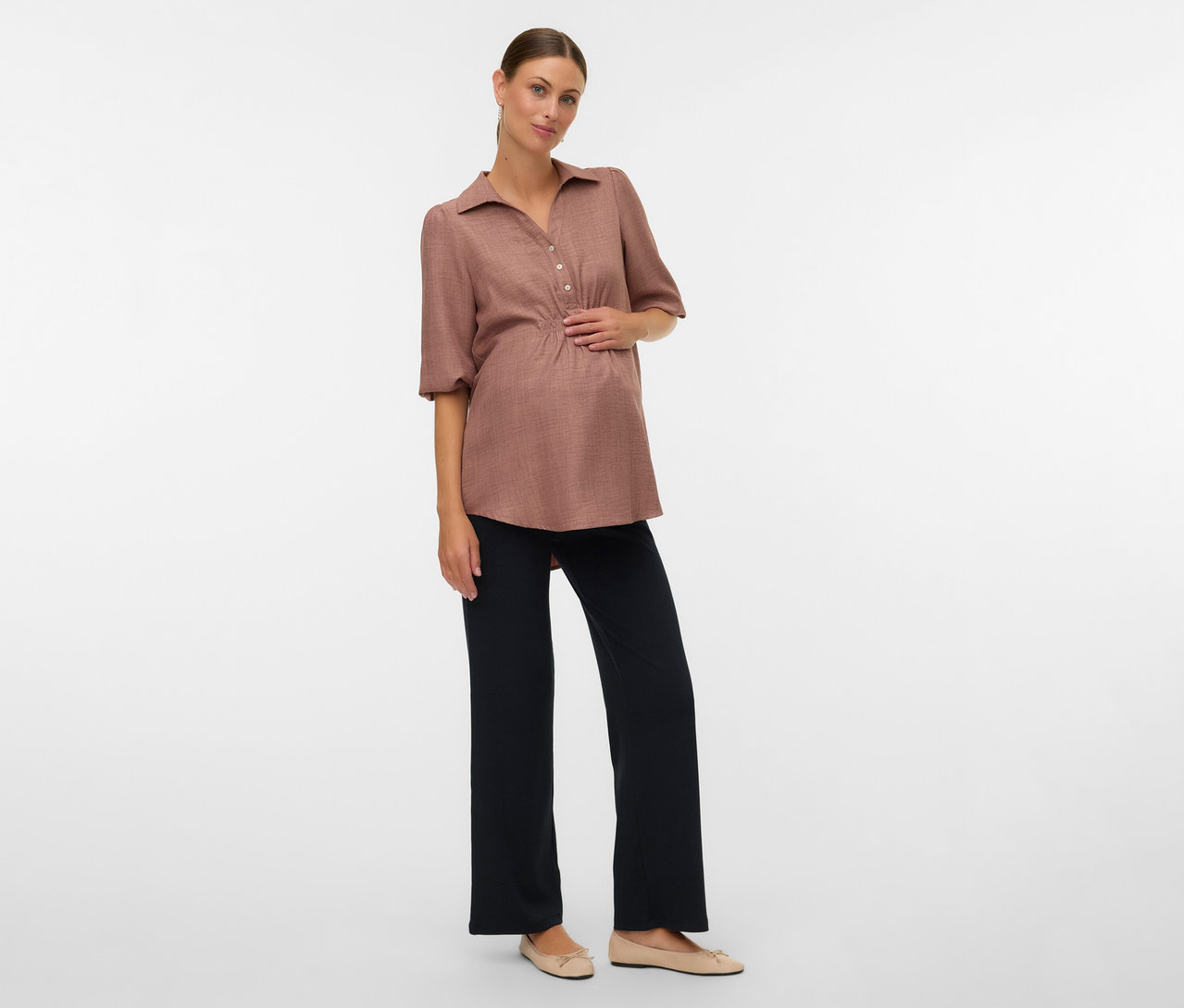 Terhes nő barna blúzban és MAMALICIOUS kismama leggingsben pózol fehér háttér előtt.