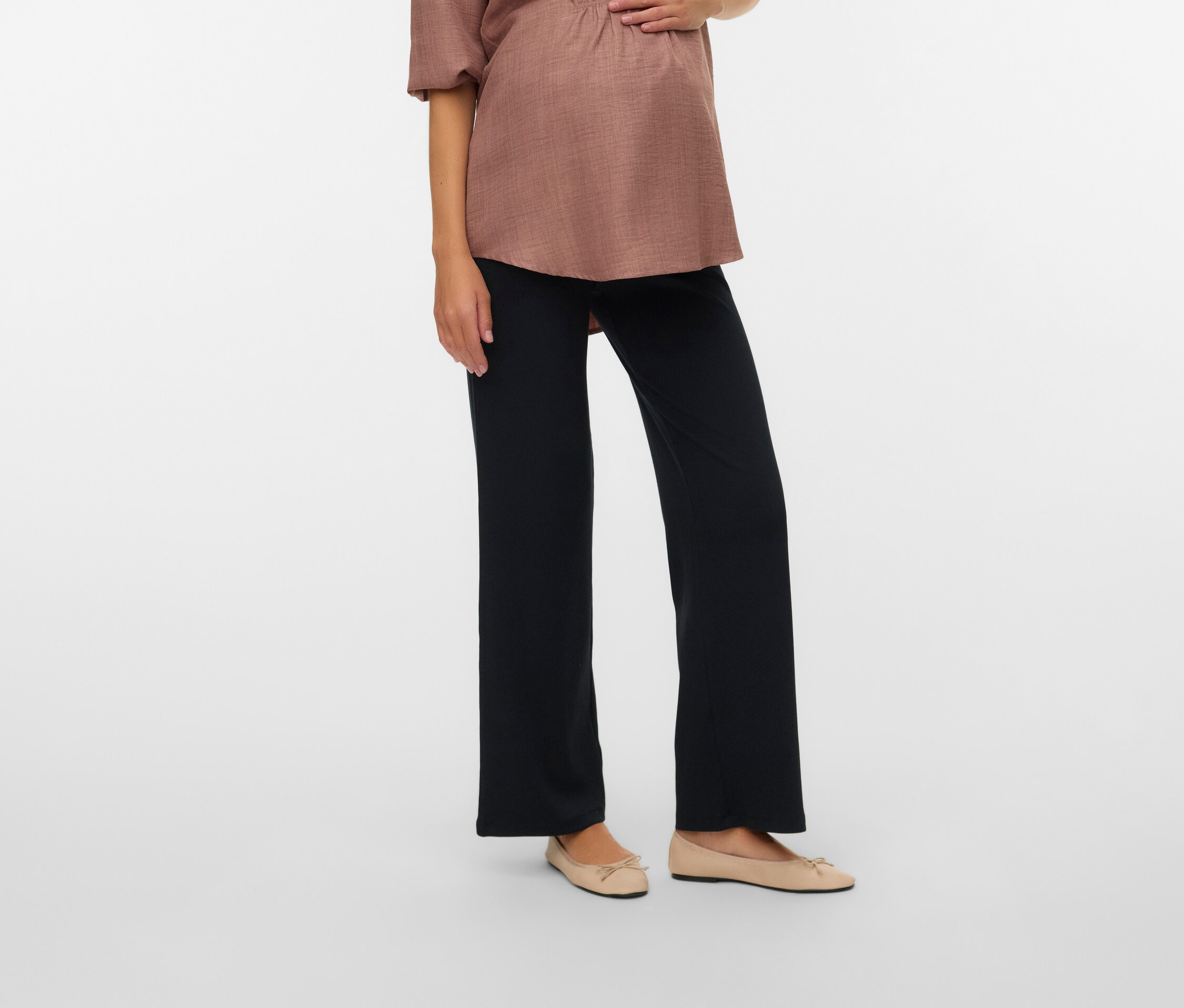 Terhes nő világosbarna blúzban és fekete nadrágban, a kezét a hasára teszi.