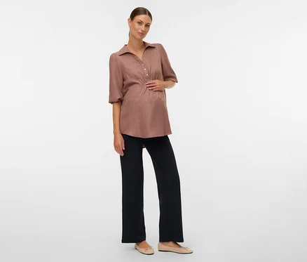 Terhes nő barna blúzban és MAMALICIOUS kismama leggingsben pózol fehér háttér előtt.