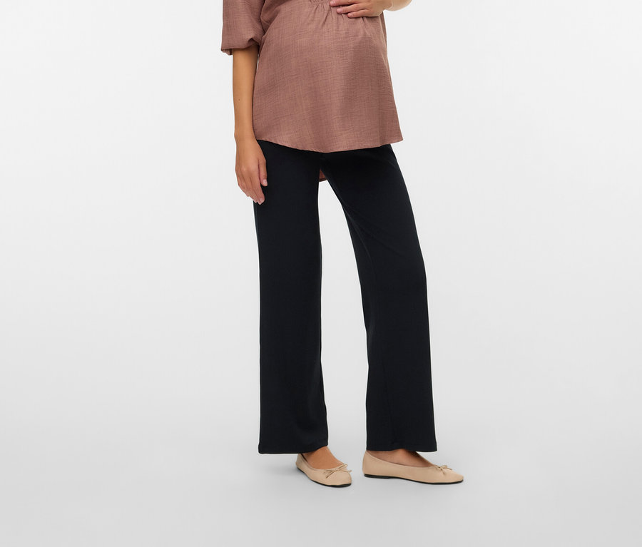 Terhes nő világosbarna blúzban és fekete nadrágban, a kezét a hasára teszi.