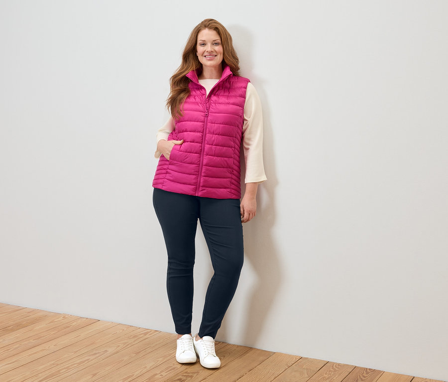 Nő rózsaszín steppelt mellényben, bézs blúzban, kék farmerben és fehér tornacipőben áll egy fehér fal mellett.