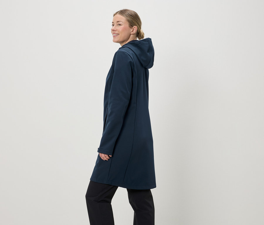 A lófarokba kötött hajú nő sötétkék, kapucnis softshell kabátot és fekete nadrágot visel világos háttér előtt.