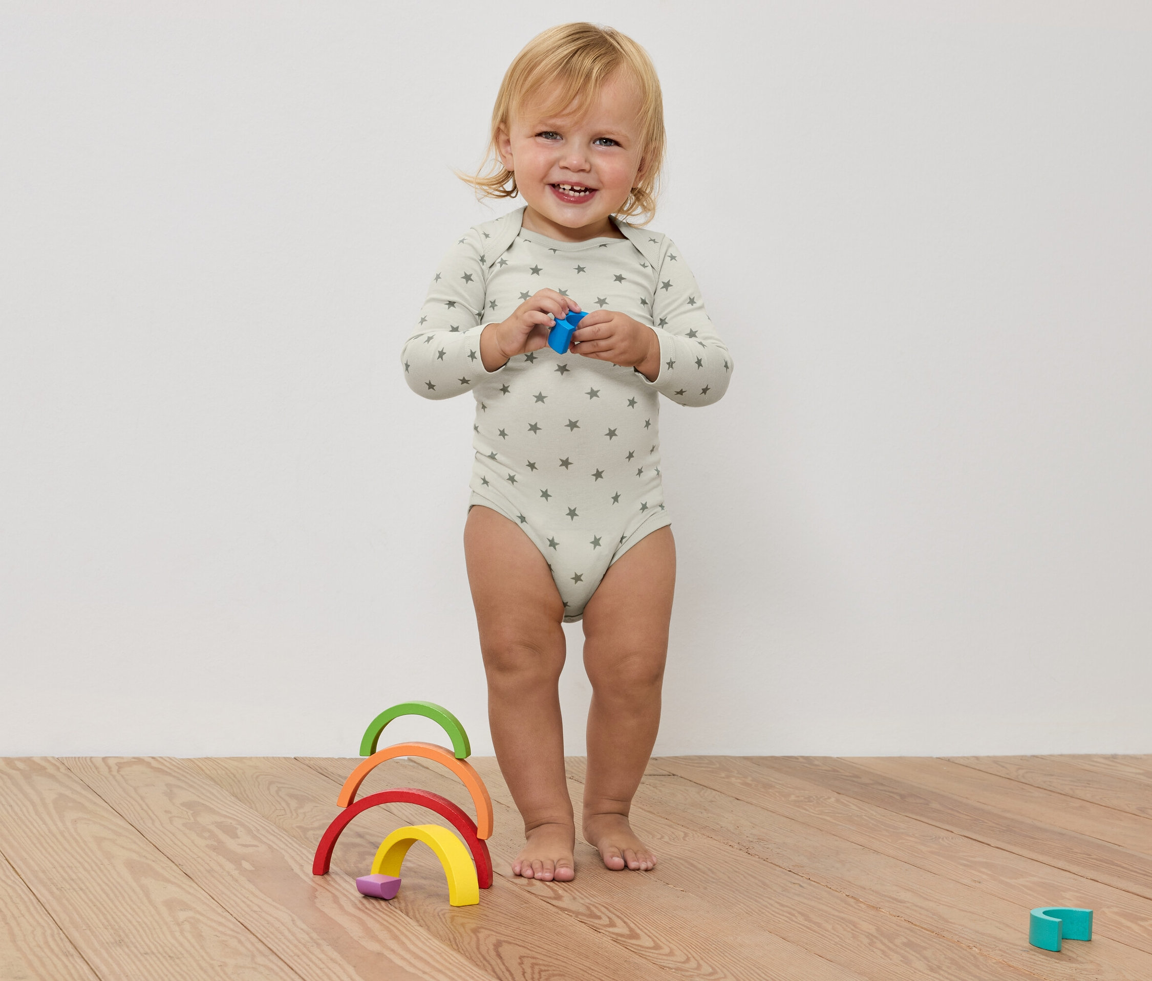 Egy mosolygó kisgyermek csillagos bodyban egy játékot tart a kezében, egy színes fa szivárvány előtt, egy fapadlón.