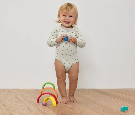 Egy mosolygó kisgyermek csillagos bodyban egy játékot tart a kezében, egy színes fa szivárvány előtt, egy fapadlón.