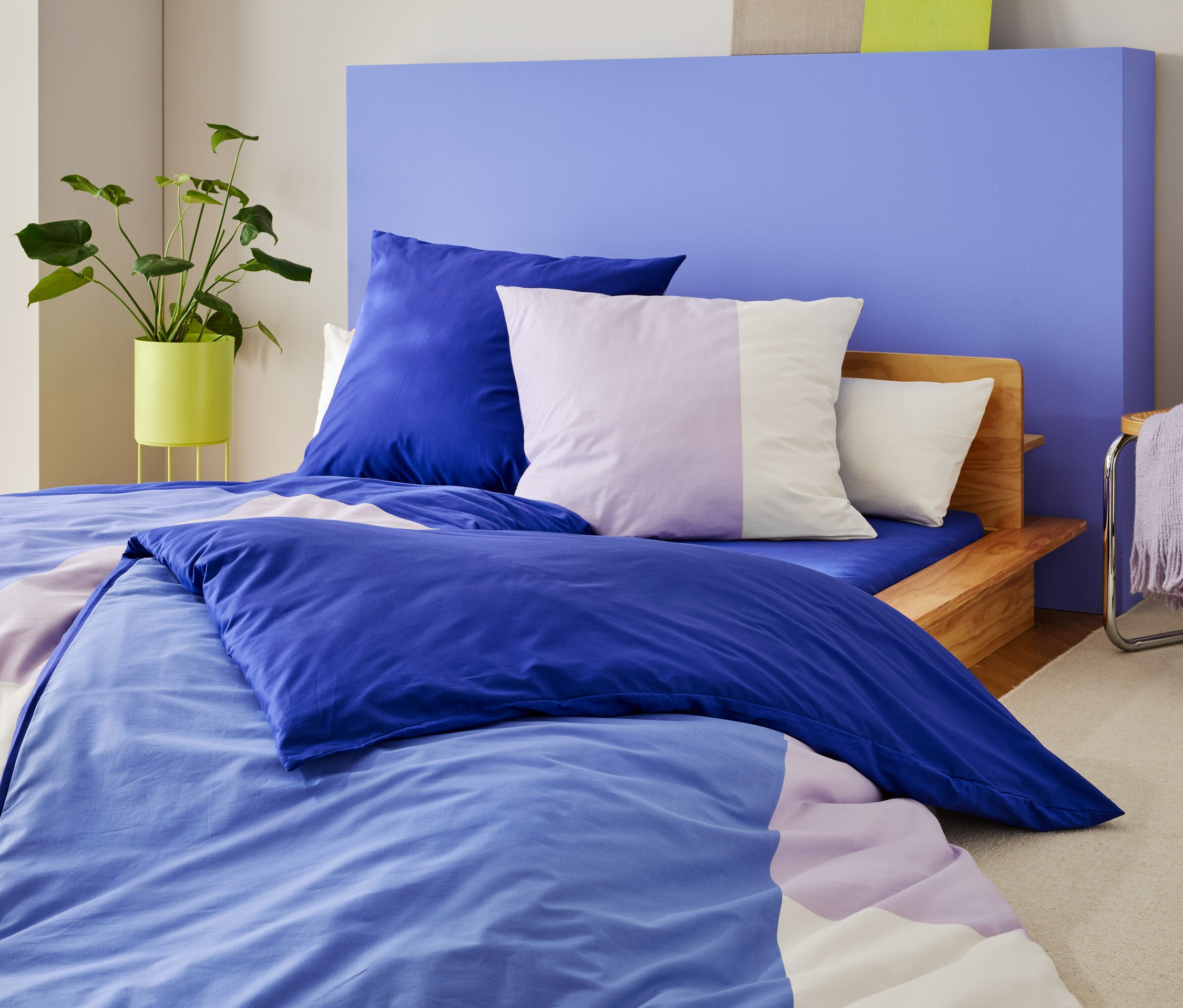 Egy ágy kék-lila ágyneművel és párnákkal egy kék fal előtt áll, mellette egy növény és egy szék egy takaróval.