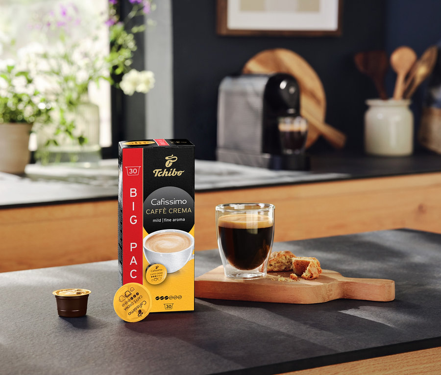 Cafissimo Caffè Crema mild kapszulák csomagja, egy csésze kávé és egy kávéfőző a konyhapulton.
