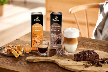 Espresso, latte macchiato, sütemény és Cafissimo Flavoured Espresso – Double Choc kapszulák egy fa asztalon.
