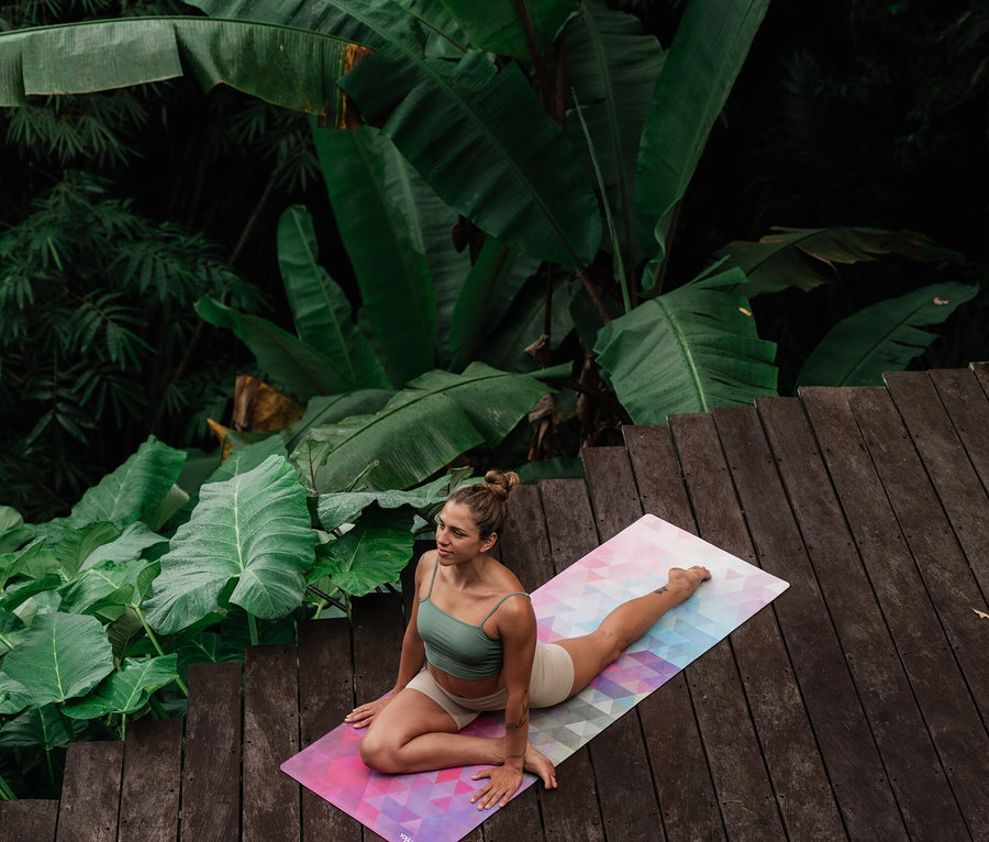 Nő galambpózban egy színes Yoga Design Lab Travel Combo jógamatracon »Tribeca«, 3,5 mm, egy trópusi növényekkel körülvett fa teraszon.