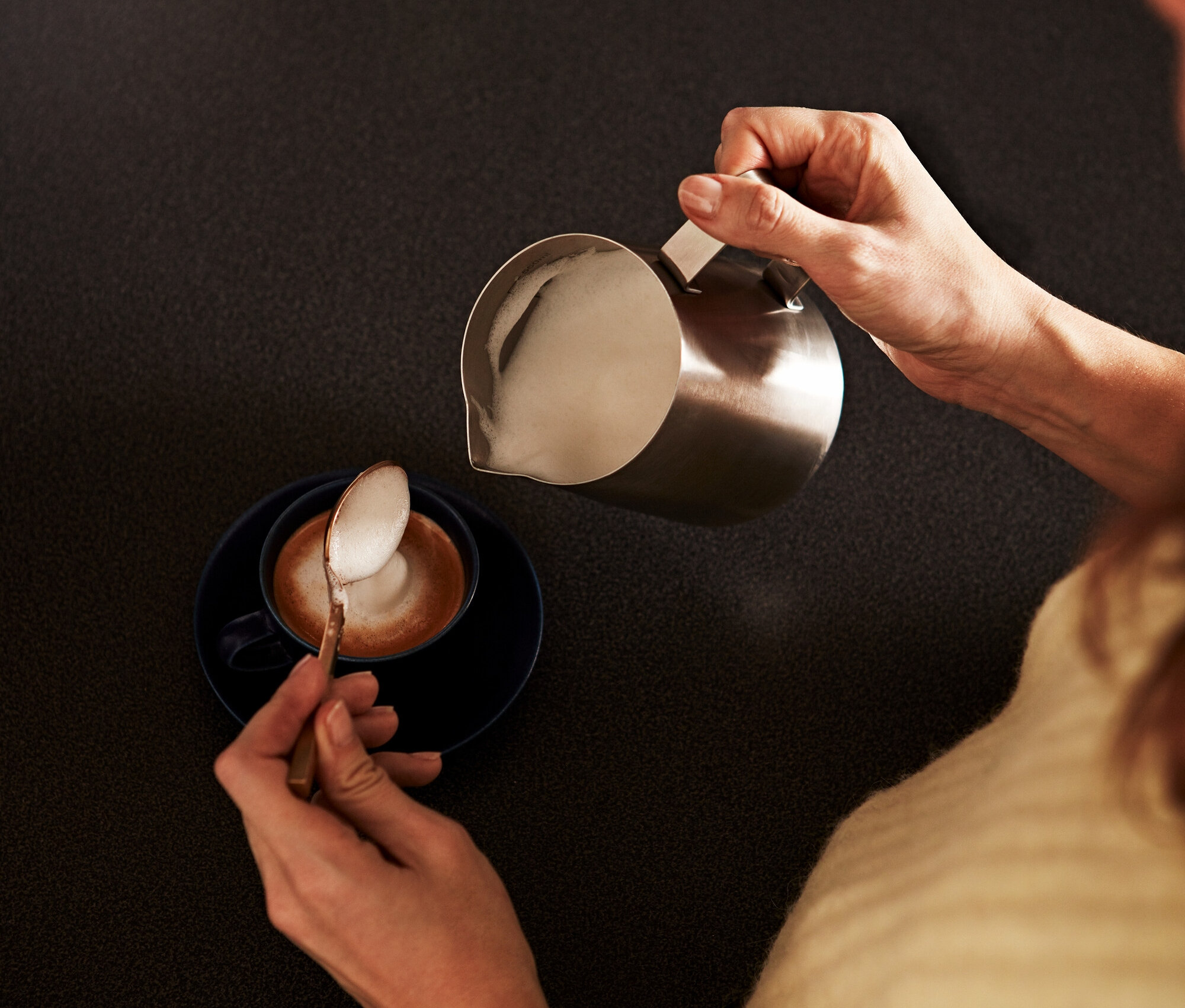 Nő önti a habosított tejet egy kis, nemesacél tejeskancsóból egy kávéscsészébe, és kanállal szedi a habot.