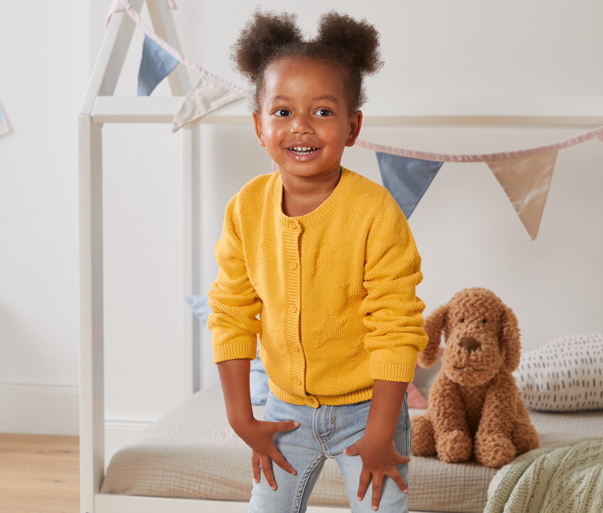 Mosolygó kislány sárga kötött kardigánban áll a gyerekszobában.