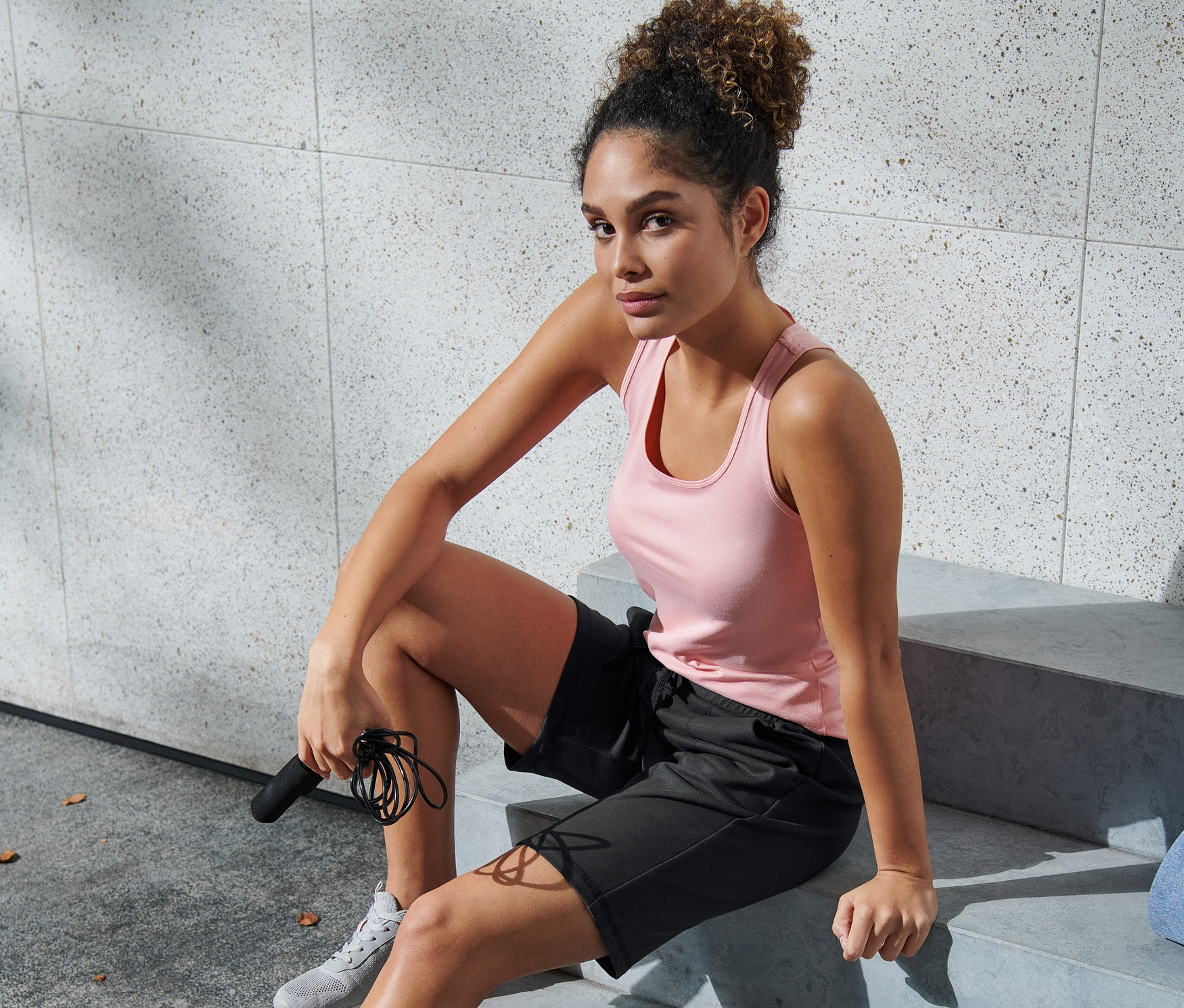 Nő ül a lépcsőn barna-fekete sport rövidnadrágban, ugrókötelet tartva.