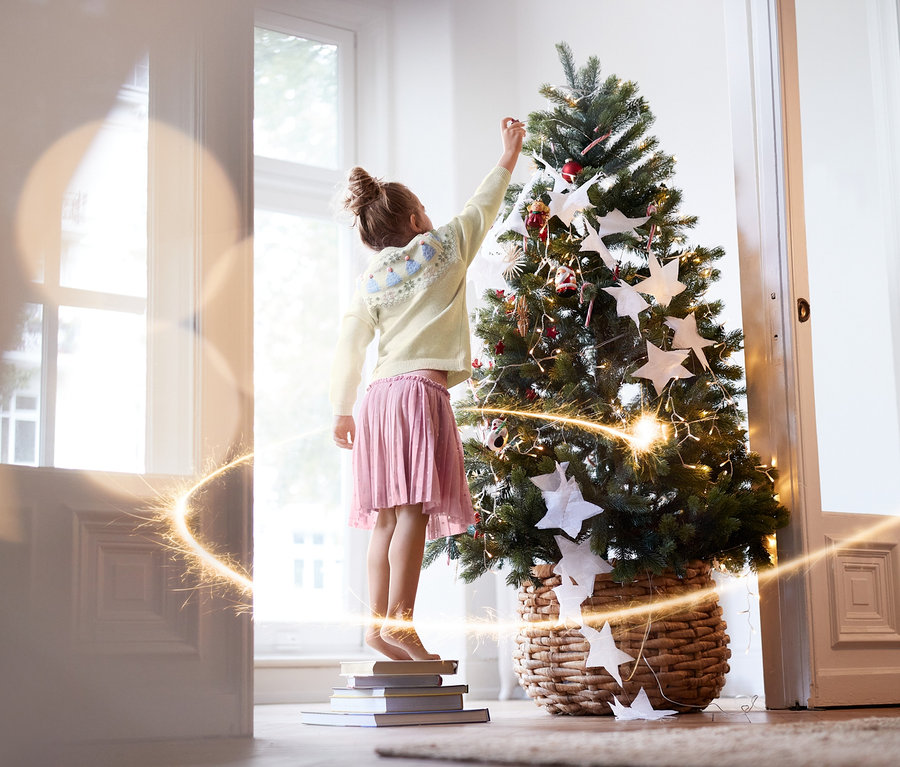 Lány áll könyveken és díszít egy karácsonyfát. Kislány tüllszoknya van rajta és egy krémszínű kislány kötött pulóver.