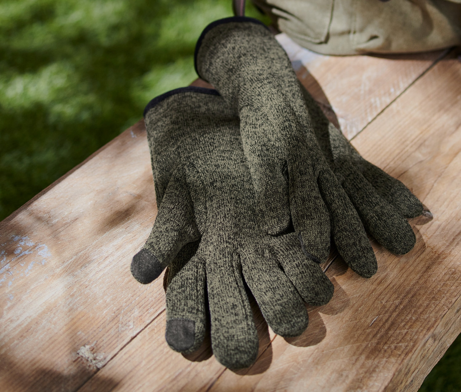 Egy pár unisex kötött polárkesztyű szürkészöld színben egy fa padon fekszik egy vászon hátizsák mellett.